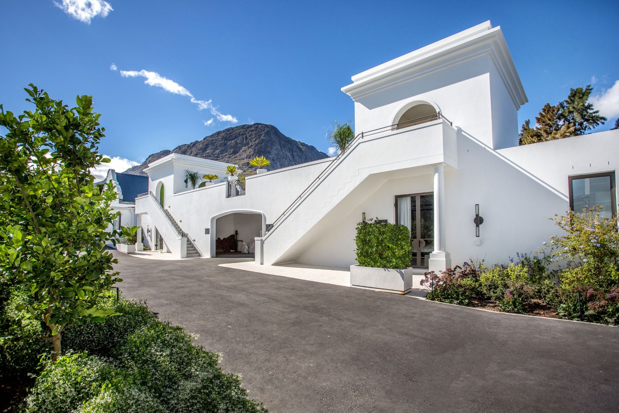 Freshly tarred driveway sweeping past a white estate building with stairs and archways, mountain behind